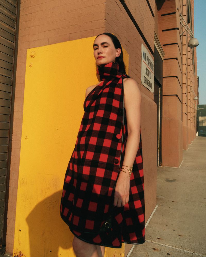 Woman in a red and black plaid dress stands outside with a brick building and yellow wall in the background, posing in sunlight.