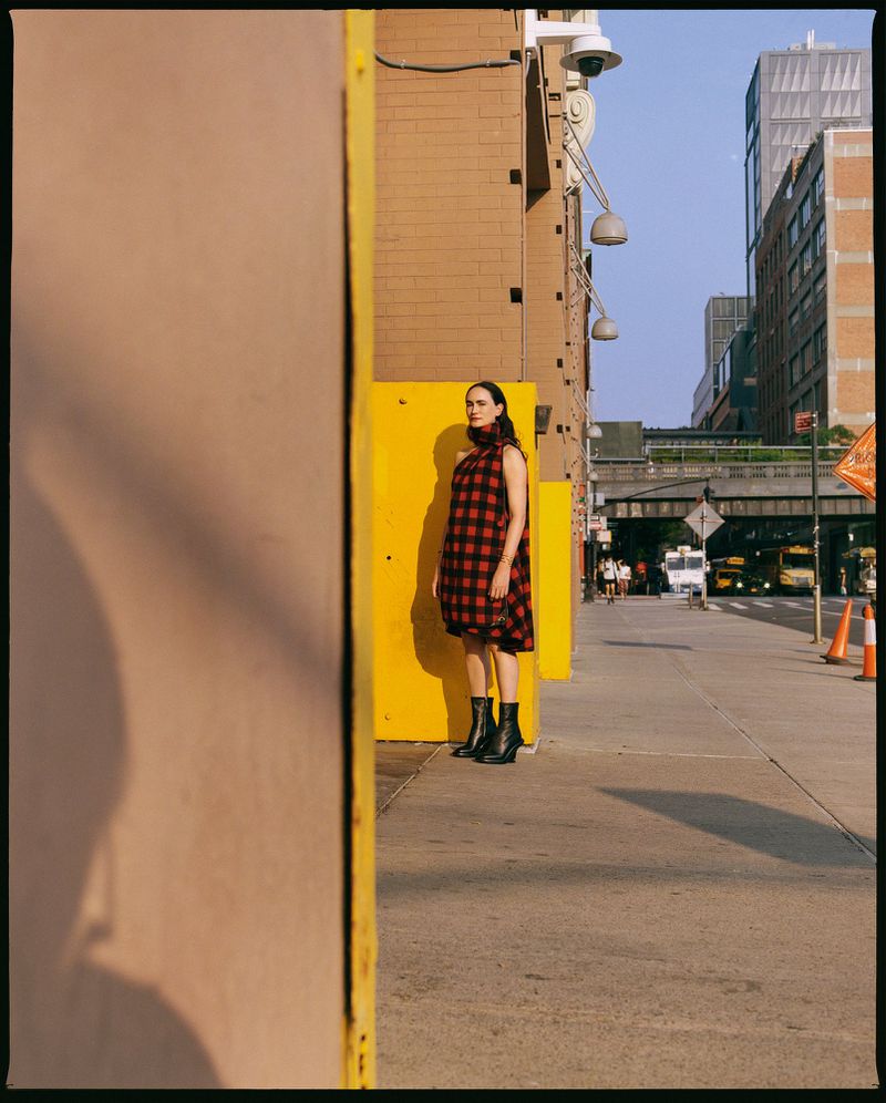 Woman in a red and black plaid dress standing on a sidewalk next to a yellow building in an urban environment, with city buildings visible.