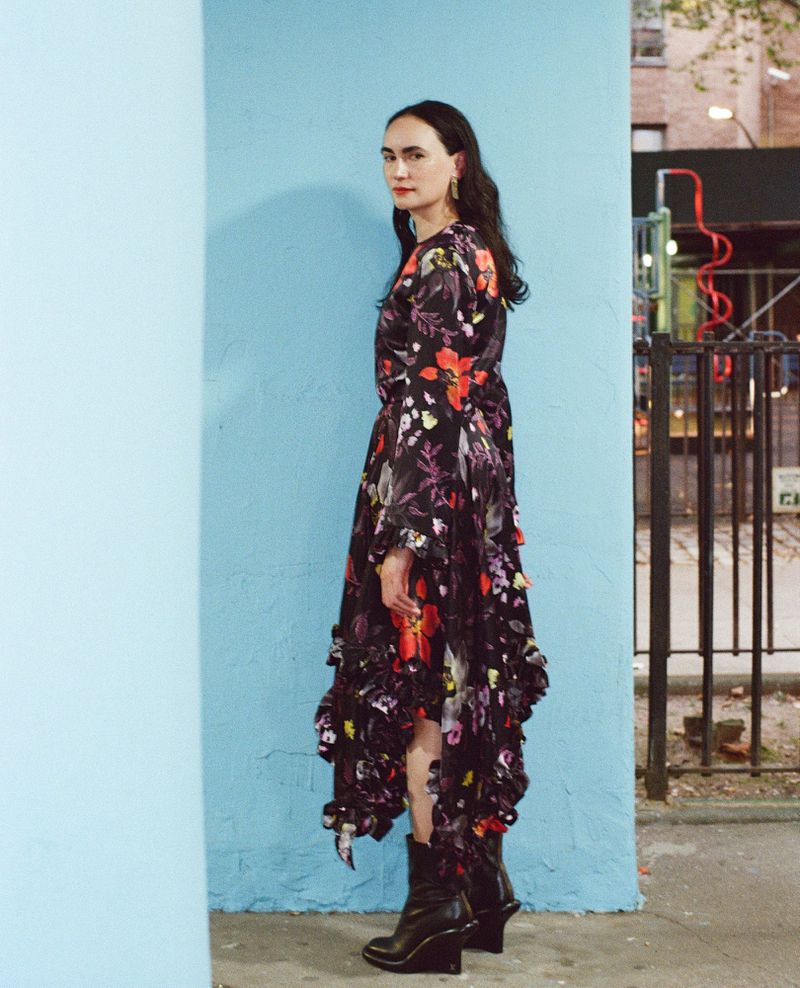 Adult woman in a floral dress and black boots poses against a light blue wall in an urban setting with a building in the background.