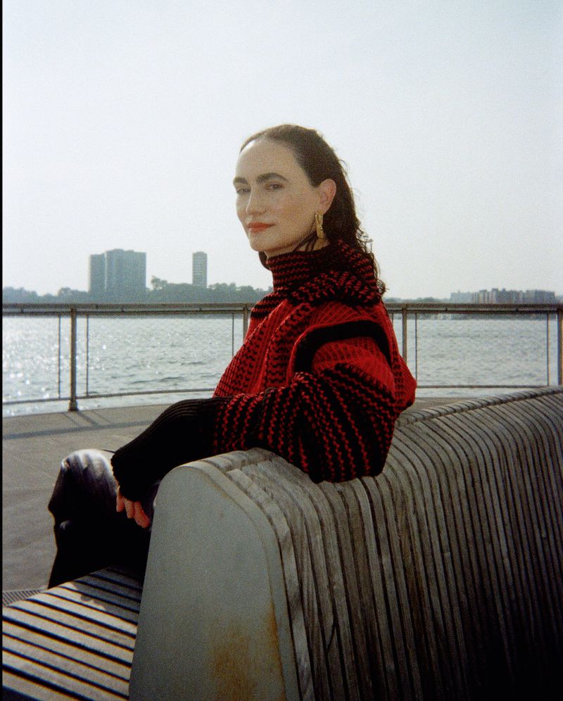 Adult woman wearing red and black striped knitwear sits on a light-colored bench near a body of water with buildings in the background.