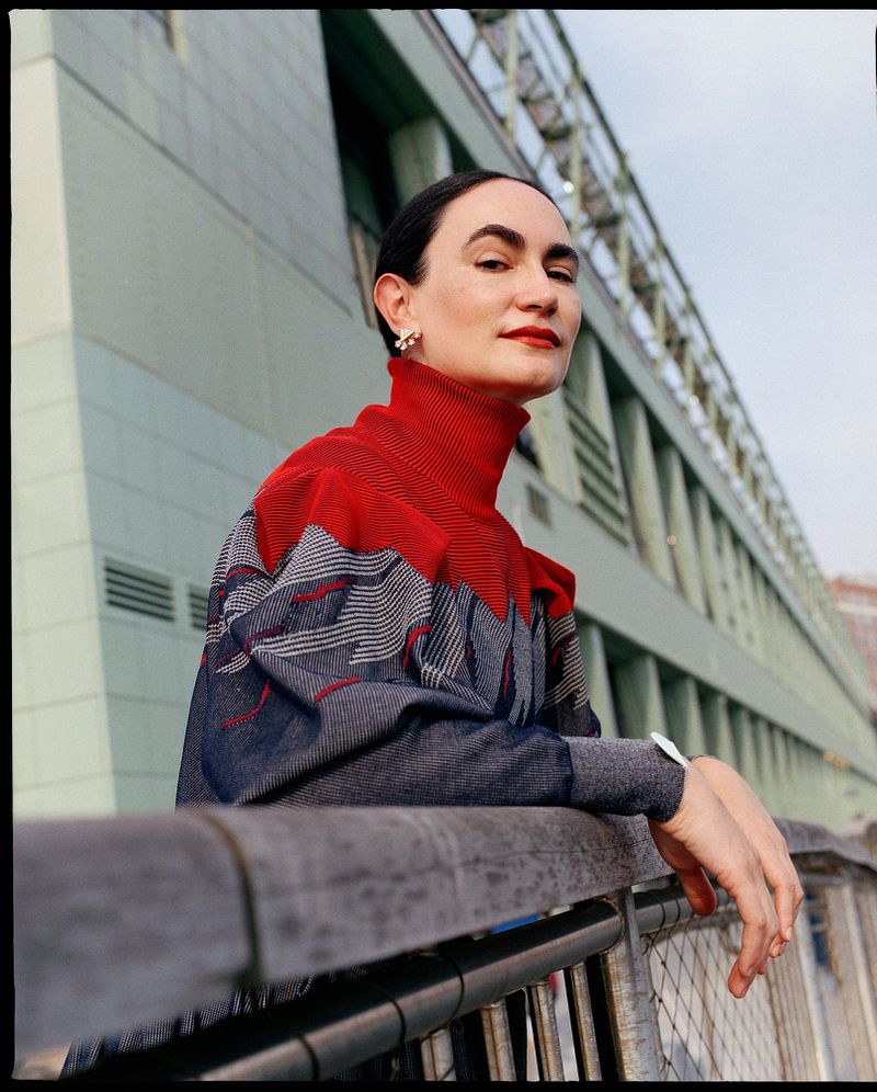 Woman with dark hair poses in red and gray patterned sweater, resting arms on rail with an industrial building in the background.