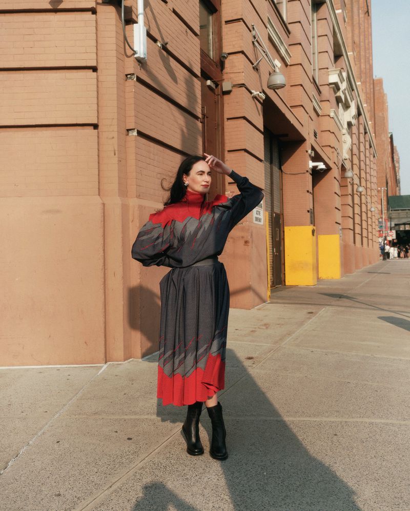 Adult woman in a red and gray dress and black boots poses on a sidewalk next to a brick building on a sunny day.