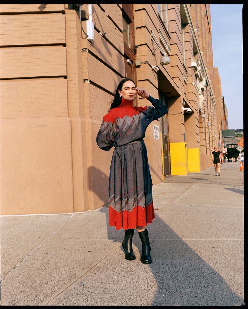 Woman in grey and red chevron dress poses on sidewalk in front of brick building on a sunny day wearing black heeled boots.