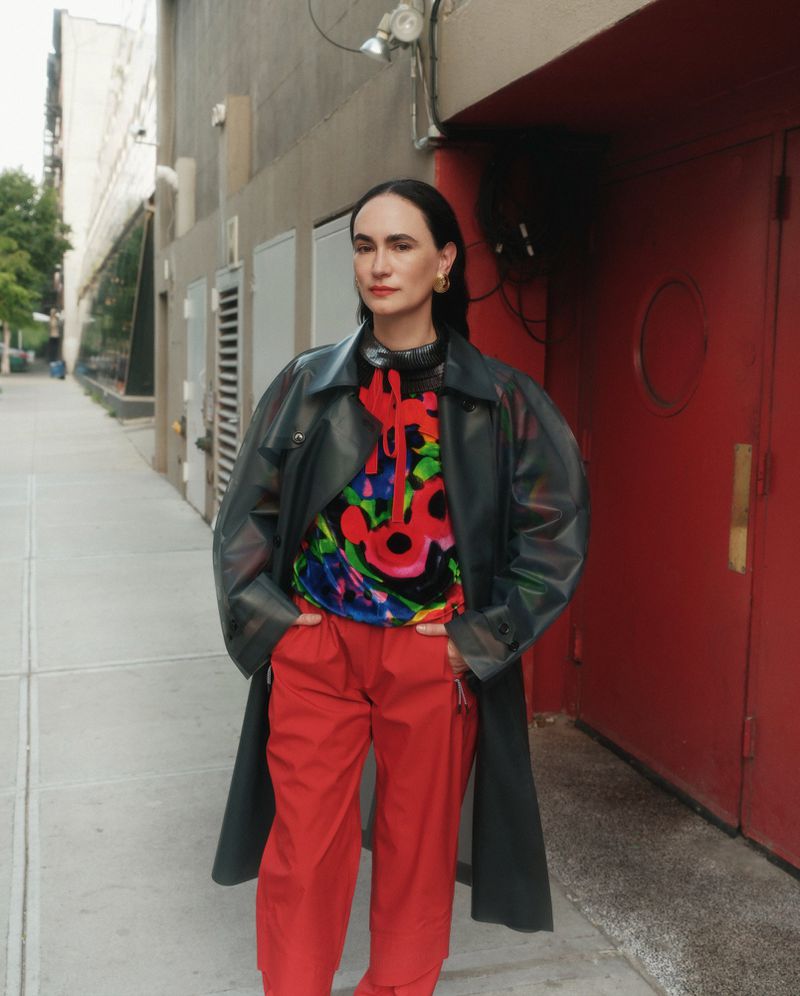Woman in a black coat and red pants poses on an urban sidewalk next to a red building with a neutral building behind her.