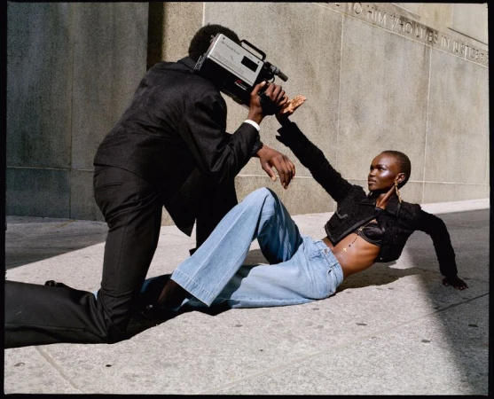 A stylish model poses with a cameraperson in an urban setting, showcasing a blend of fashion and vintage camera equipment against a building.