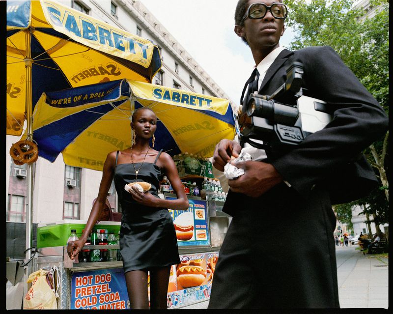 A black man films a Black woman in a dress, who holds a hotdog, at a Sabrett hotdog stand in a city with buildings and trees.