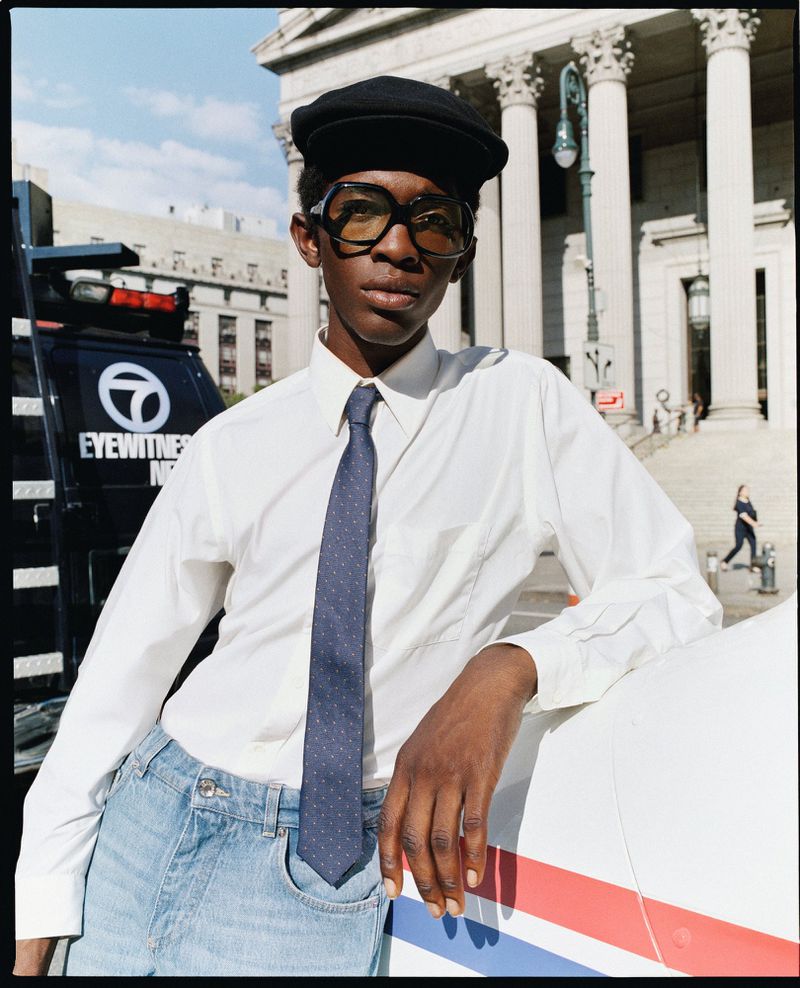 A stylish black man with glasses and a cap leans against a news van in an urban setting with a classical building in the background.
