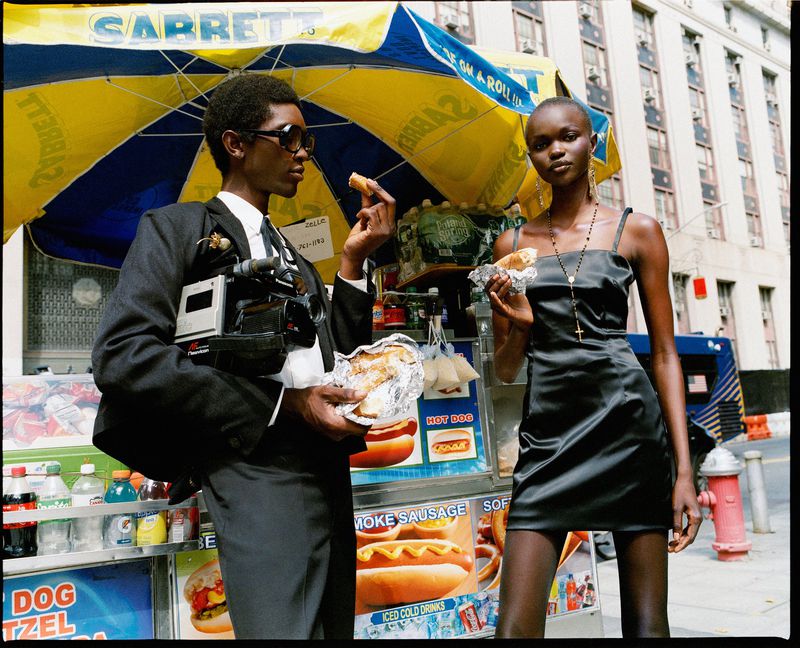 Two fashionably dressed individuals, a man and a woman, stand beside a New York City food cart, each holding a snack; urban setting.