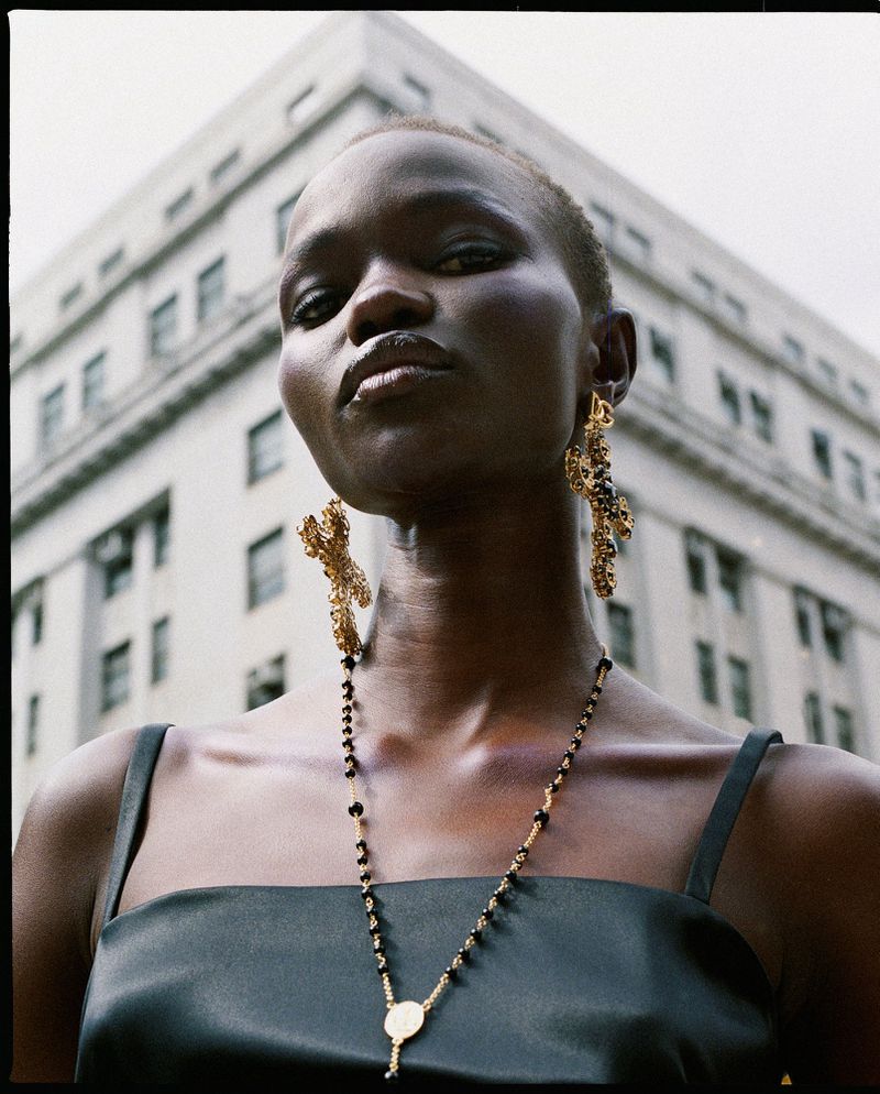 Fashion shot of an African American woman wearing gold earrings and a necklace, with a neutral facial expression against a building backdrop.