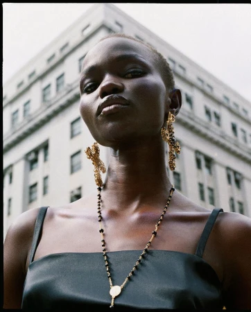 Fashion shot of an African American woman wearing gold earrings and a necklace, with a neutral facial expression against a building backdrop.