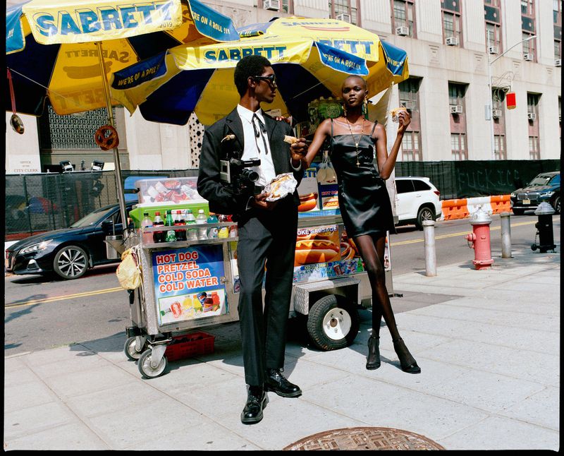 Stylish man and woman posing with a hot dog cart on a city street. Both are eating food. The man holds a camera in his left hand.