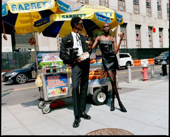 Stylish man and woman posing with a hot dog cart on a city street. Both are eating food. The man holds a camera in his left hand.