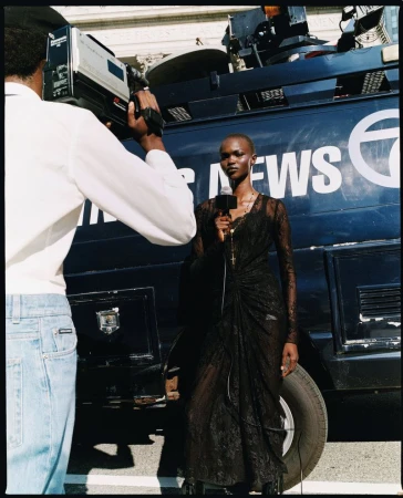 Female reporter with a microphone reporting news in front of a news van being filmed by a cameraman in an outdoor urban setting.