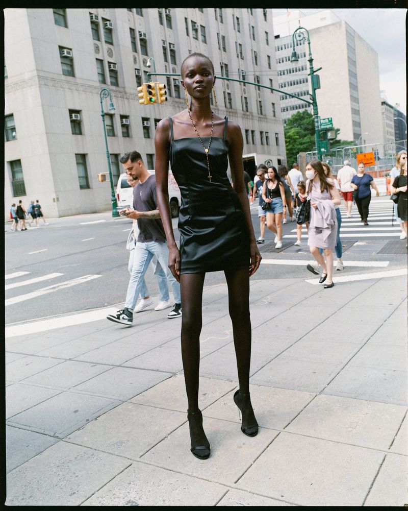 Fashion shot of a tall, slim, African-American woman in a black dress posing in the city on a sunny day, with a crowd of people.