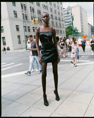 Fashion shot of a tall, slim, African-American woman in a black dress posing in the city on a sunny day, with a crowd of people.