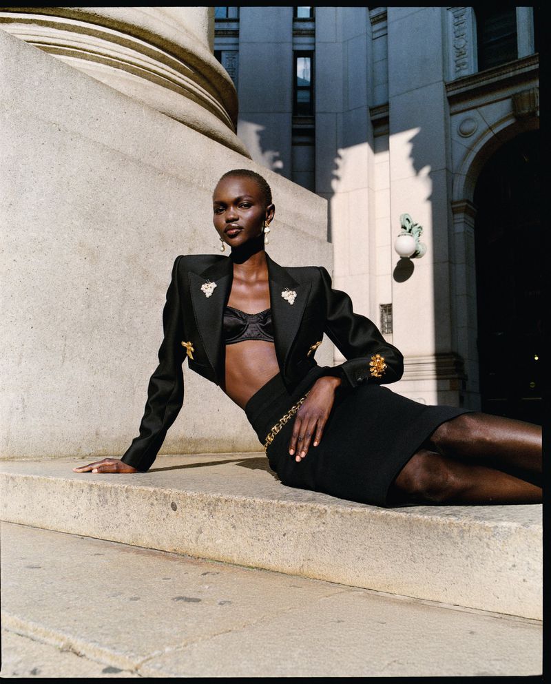 Fashion shot of a bald African-American woman in a black jacket and skirt sitting on stone steps in front of a building.