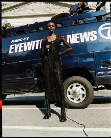A fashion model in a black lace dress stands in front of a KABC-TV news van on a city street, holding a microphone, under daylight.