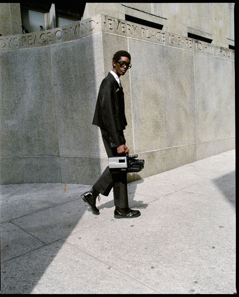 Man in black suit walks outdoors carrying retro camcorder in urban environment, with concrete wall and paving stones.