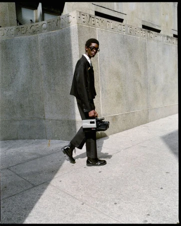 Man in black suit walks outdoors carrying retro camcorder in urban environment, with concrete wall and paving stones.