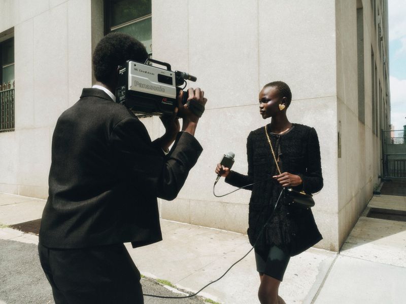Woman in black outfit with heart earrings being interviewed outdoors while a videographer records with a vintage Panasonic camera.