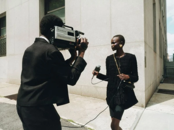 Woman in black outfit with heart earrings being interviewed outdoors while a videographer records with a vintage Panasonic camera.