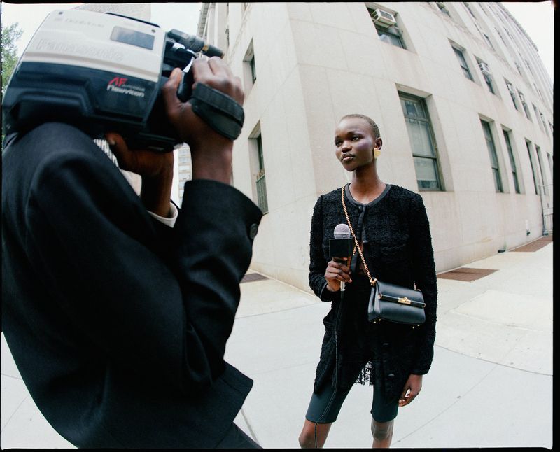 Fashion shoot of a reporter with a microphone being filmed by a cameraman outside a light-colored building, shot at eye-level, medium shot.