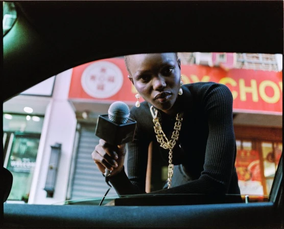 Model Adau Aguer leans toward a car window holding a reporter microphone, wearing a black ribbed sweater and chunky gold chain necklace.