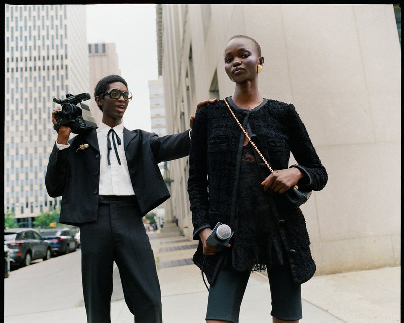 Two African models pose on a city sidewalk; one holds a vintage video camera and the other has a microphone. Fashionable editorial shot.