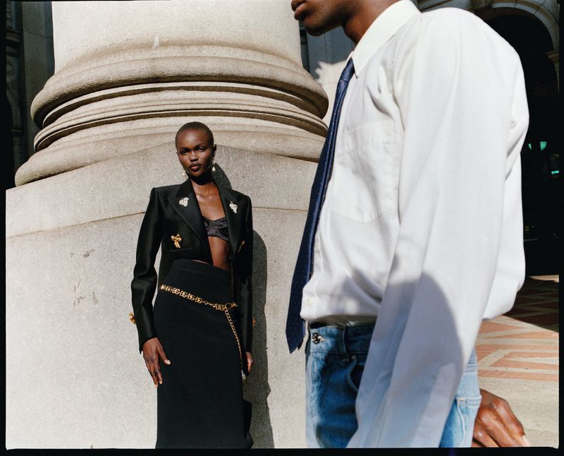 Fashion shot with a Black woman in a blazer and skirt standing next to a man in a dress shirt and jeans by a stone column.