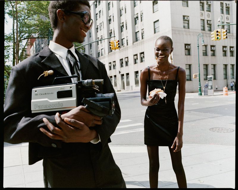 Fashion photo of two people, one in a suit holding a video camera and the other in a black dress eating a sandwich on a city street.
