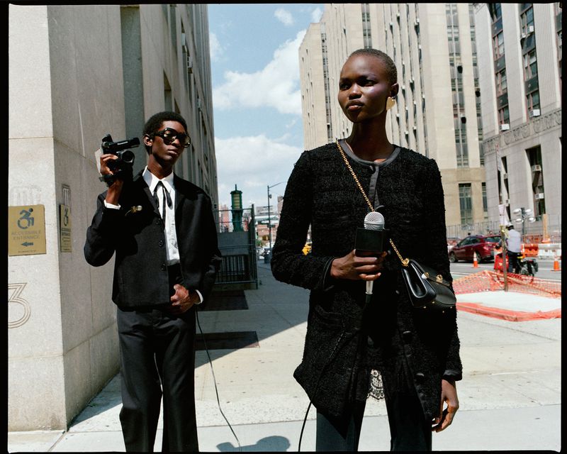 Two fashion models stand on a city street. An adult male in a black suit holds a camera; an adult female holds a microphone.