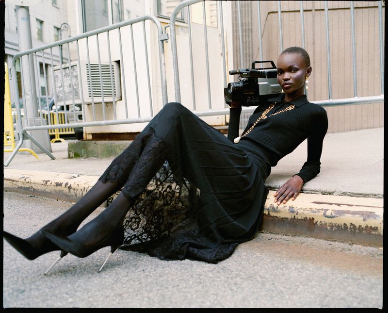 Woman in black poses on street with vintage video camera. She's wearing dark, formal attire with lace details and pointed heels.