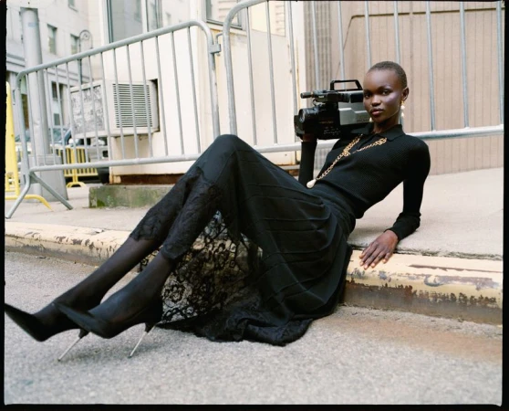 Woman in black poses on street with vintage video camera. She's wearing dark, formal attire with lace details and pointed heels.