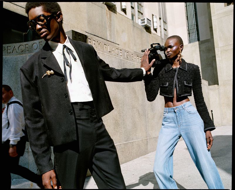 Two fashion models pose on a city street; a man in a suit stands beside a woman holding a video camera, near a stone building.
