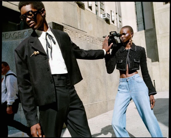 Two fashion models pose on a city street; a man in a suit stands beside a woman holding a video camera, near a stone building.