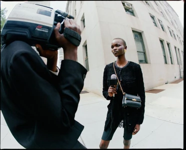 Black model holding a microphone while being filmed by a camera operator on a city sidewalk