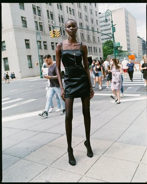 Model Adau Aguer stands on an urban sidewalk wearing a black satin mini dress, cross necklace, dark tights, and strappy heels.