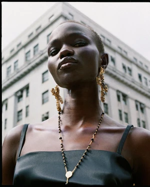 Model Adau Aguer wearing ornate gold earrings and a beaded necklace against a tall stone building exterior.