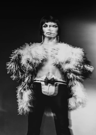 Monochrome portrait of a gender-ambiguous human in a feather boa, dress shirt with bow tie, and dark pants against a dark background.