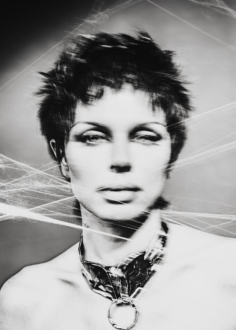 Monochrome portrait of a person with short spiky hair and a large metal necklace, styled with a web-like effect. Fashionable, striking.