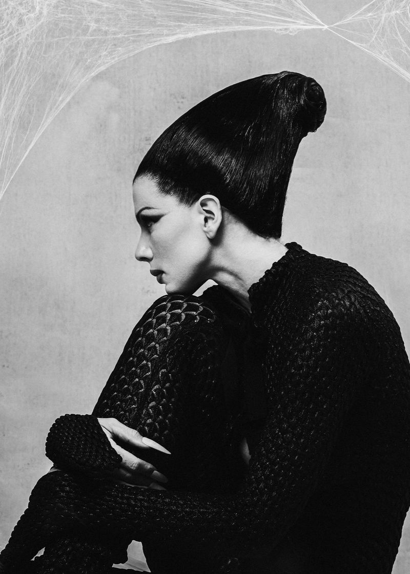 Woman in black textured jumpsuit with striking hairstyle poses in a studio against a web background in this black and white fashion portrait.