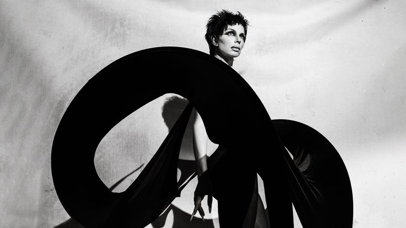 Black and white portrait of a person with short hair wearing black fabric, posing in front of a textured white background.