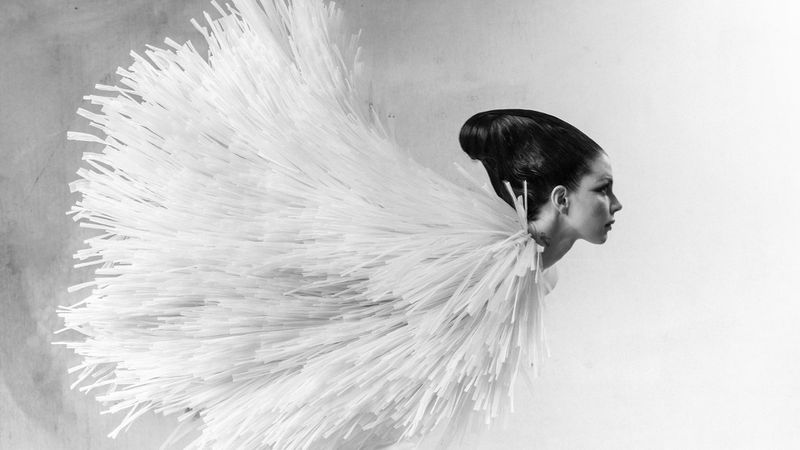 Profile shot of woman with dark hair styled in a bun, wearing a white textured garment, set against a monochromatic grey backdrop.