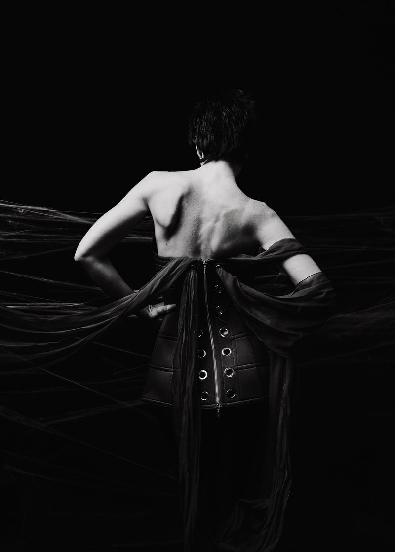 Back view of adult female in corset and draped fabric against a black background in a studio shot showcasing fashion elements.