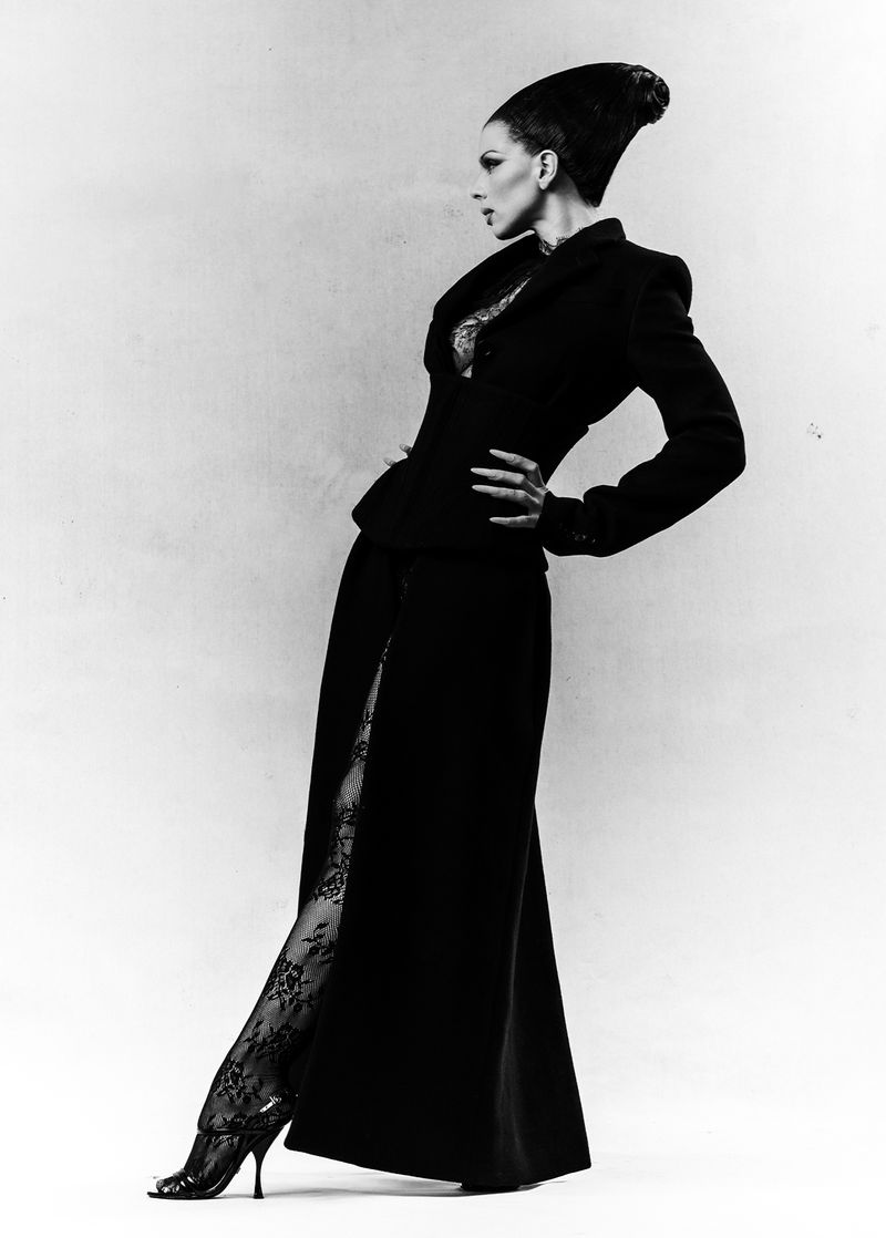 Fashionable woman in black jacket and skirt with lace stockings poses against a grey backdrop in a studio setting. Side view.