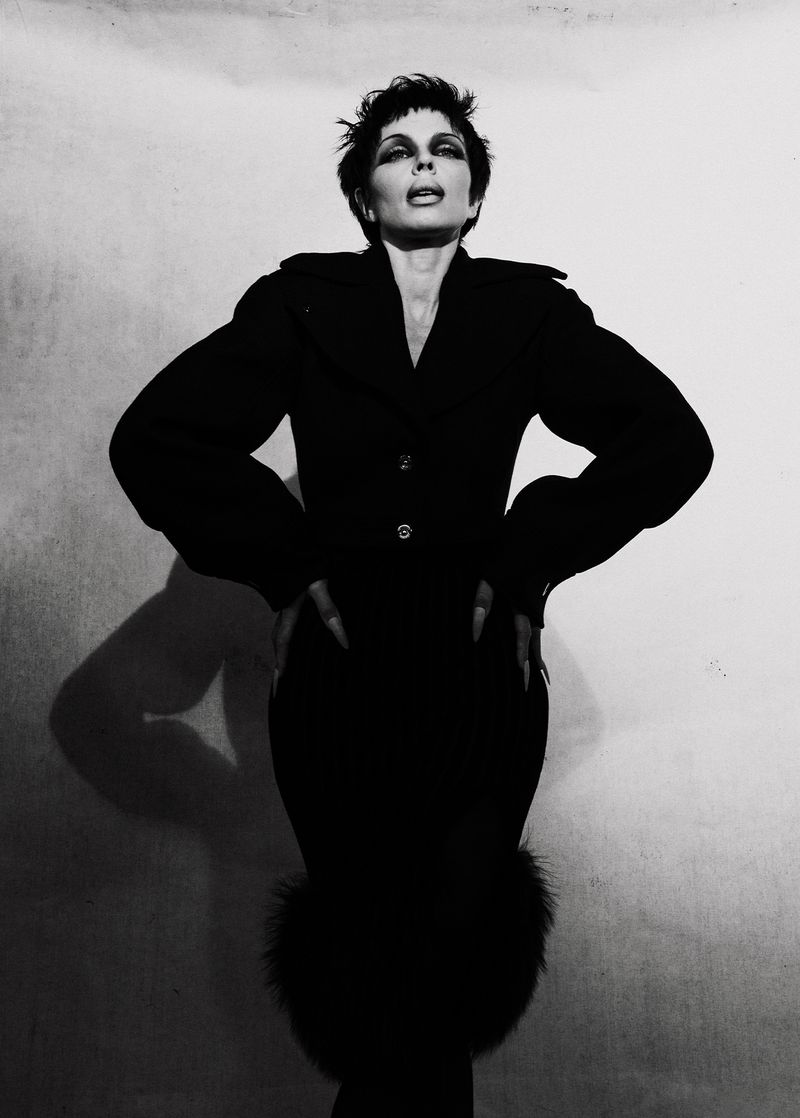 Monochrome portrait of a woman in a dark coat with fur trim, hands on hips, against a mottled grey background with dramatic lighting.