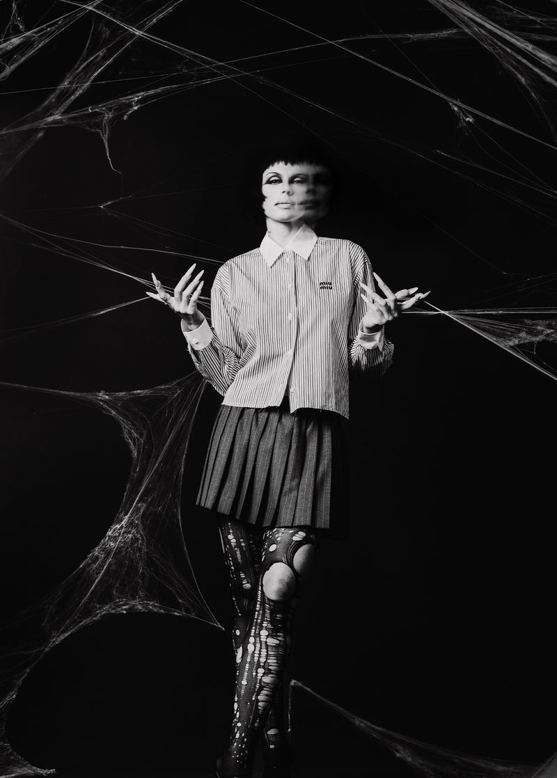 Monochrome studio shot of a model in striped shirt, pleated skirt, and torn tights, against a spiderweb-decorated dark backdrop.