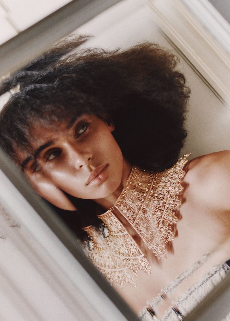 Glamour portrait of a woman with dark curly hair, wearing an elaborate lace collar with gold beads, leaning her head on her hand.