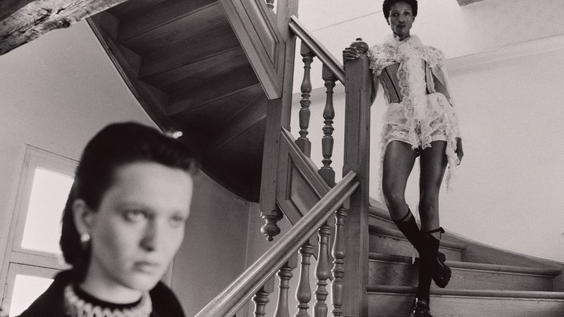 Black-and-white fashion shot of two women indoors: one on a wooden staircase wearing a lace outfit, the other blurred.