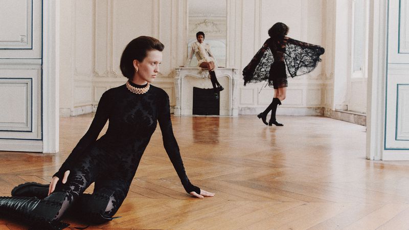 Three women model vintage-inspired fashion in an elegant interior. One sits on the floor, one on a mantel, and the third stands.
